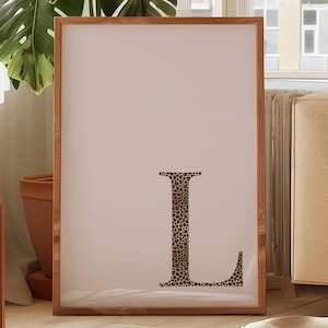 Könnte beinhalten: Ein gerahmter Kunstdruck mit einem großen Buchstaben "L" in Leopardenmuster. Das Kunstwerk ist vor einem hellen Hintergrund platziert und in Naturholz gerahmt. Der Druck wird in einem hellen, sonnendurchfluteten Raum ausgestellt.