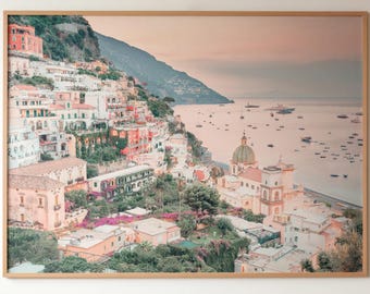 May include: A large, square, framed wall art print featuring a coastal landscape of Positano, Italy. The image displays multi-colored buildings built into a steep hillside, lush green vegetation, a sandy beach, and boats on the calm sea at sunset.