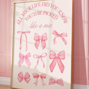 May include: A framed print featuring various pink watercolor bows. The text "ALL YOUR LIFE DID YOU KNOW YOU'D BE PICKED like a rose" is displayed in pink script. The artwork is set against a light background and framed in gold.