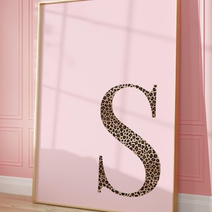 May include: A framed pink art print featuring a large letter "S" in a leopard print pattern. The artwork is displayed against a pink wall with molding, and the frame has a gold finish. The print leans against the wall on a wooden floor.