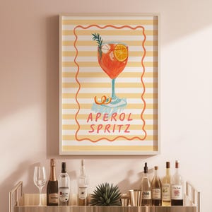 May include: An Aperol Spritz cocktail art print displayed on a wall. The artwork features a watercolor illustration of the cocktail in a tall glass with an orange slice and rosemary sprig. The background is a yellow and white striped pattern with the words 'APEROL SPRITZ' in the center. A bar cart with bottles and glasses is in the foreground.