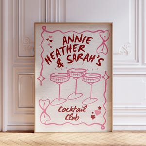 May include: A framed print with a light beige background and pink illustrations. The text reads "Annie, Heather & Sarah's Cocktail Club" above three pink champagne glasses. The frame is a light brown color.