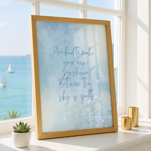 Könnte beinhalten: Ein gerahmter Druck mit hellblauem Aquarellhintergrund und dem Zitat "You had to make your own sunshine, But now the sky is opatte" in blauer Schrift. Der Rahmen hat eine helle Holzfarbe. Zwei goldene Kerzenhalter und eine kleine Sukkulente in einem weißen Topf stehen auf der Fensterbank.