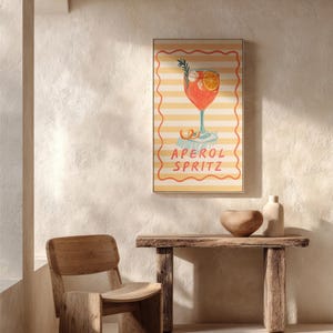 K&ouml;nnte beinhalten: Ein gerahmter Druck mit einer Illustration eines Aperol Spritz Cocktails. Das Kunstwerk zeigt einen leuchtend orangefarbenen Cocktail mit Rosmarin und einer Orangenscheibe auf gestreiftem Hintergrund. Die Worte "APEROL SPRITZ" sind darunter geschrieben.