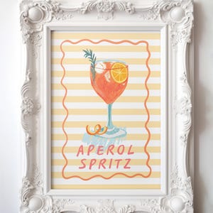 K&ouml;nnte beinhalten: Ein gerahmter Druck mit einer Illustration eines Aperol Spritz Cocktails. Das Kunstwerk zeigt einen leuchtend orangefarbenen Cocktail mit einer Orangenscheibe und einem Rosmarinzweig, vor einem gelb-wei&szlig; gestreiften Hintergrund. Die Worte "APEROL SPRITZ" sind in Rosa geschrieben.