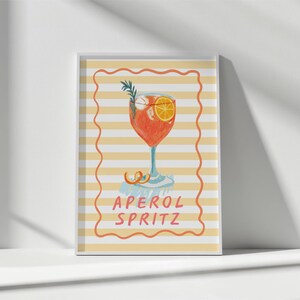 K&ouml;nnte beinhalten: Ein gerahmter Druck mit einer Illustration eines Aperol Spritz Cocktails. Das Getr&auml;nk befindet sich in einem Stielglas mit einer Orangenscheibe und Rosmarinzweig. Der Hintergrund hat gelbe und wei&szlig;e Streifen, mit den Worten "APEROL SPRITZ" in einem roten Rahmen.
