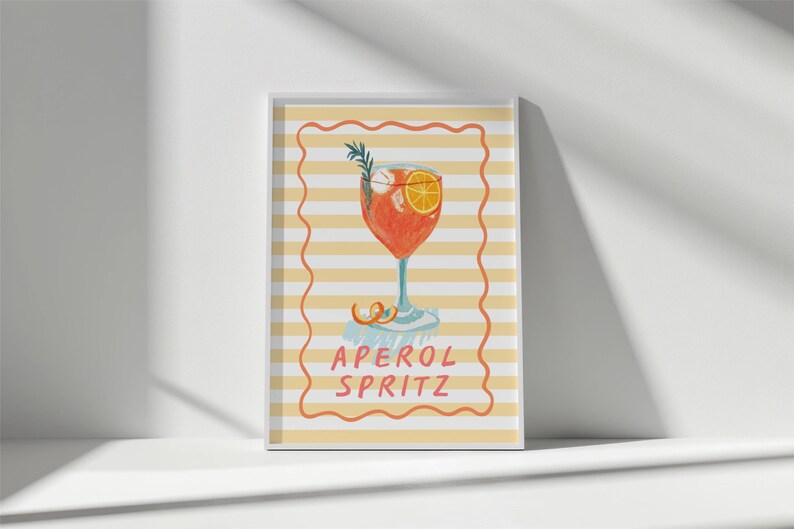 K&ouml;nnte beinhalten: Ein gerahmter Druck mit einer Illustration eines Aperol Spritz Cocktails. Das Getr&auml;nk befindet sich in einem Stielglas mit einer Orangenscheibe und Rosmarinzweig. Der Hintergrund hat gelbe und wei&szlig;e Streifen, mit den Worten "APEROL SPRITZ" in einem roten Rahmen.