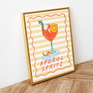 K&ouml;nnte beinhalten: Gerahmter Kunstdruck mit einer Illustration eines Aperol Spritz Cocktails. Das Getr&auml;nk ist in einem hohen Glas mit einer Orangenscheibe und Rosmarinzweig. Der Hintergrund hat gelbe und wei&szlig;e Streifen, darunter steht der Text "APEROL SPRITZ".