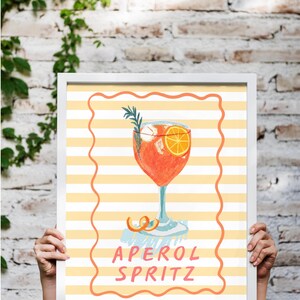 K&ouml;nnte beinhalten: Gerahmter Kunstdruck eines Aperol Spritz Cocktails. Die Aquarellillustration zeigt einen leuchtend orangefarbenen Cocktail mit einer Orangenscheibe und Rosmarinzweig. Der Hintergrund hat gelbe und wei&szlig;e Streifen mit dem Text 'APEROL SPRITZ'.