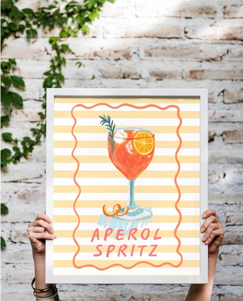 K&ouml;nnte beinhalten: Gerahmter Kunstdruck eines Aperol Spritz Cocktails. Die Aquarellillustration zeigt einen leuchtend orangefarbenen Cocktail mit einer Orangenscheibe und Rosmarinzweig. Der Hintergrund hat gelbe und wei&szlig;e Streifen mit dem Text 'APEROL SPRITZ'.