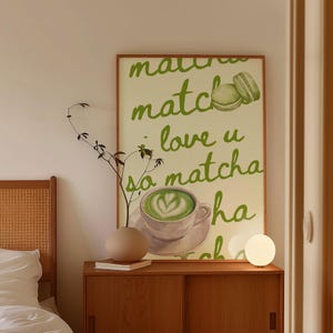May include: A framed poster with the words "matcha, love u so matcha" in green script. The poster features a watercolor illustration of a matcha latte and a macaron. A small, round, white lamp sits on a wooden cabinet.