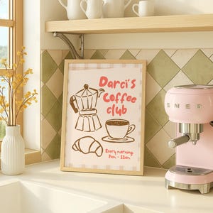 May include: A framed sign with the text "Darci's Coffee Club" and illustrations of a coffee maker, cup, and croissant. A pink espresso machine is on the right. A white vase with yellow flowers is on the left.