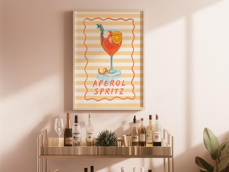 May include: An Aperol Spritz cocktail art print displayed on a wall. The artwork features a watercolor illustration of the cocktail in a tall glass with an orange slice and rosemary sprig. The background is a yellow and white striped pattern with the words 'APEROL SPRITZ' in the center. A bar cart with bottles and glasses is in the foreground.