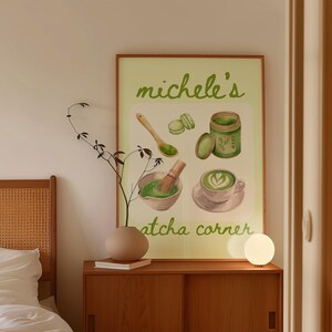 Peut inclure: Une affiche encadr&eacute;e avec le texte "michele's atcha corner" pr&eacute;sente des illustrations &agrave; l'aquarelle de la pr&eacute;paration du th&eacute; matcha : un bol, un fouet, une cuill&egrave;re, des macarons et une tasse de latte matcha. L'affiche est sur un meuble en bois.