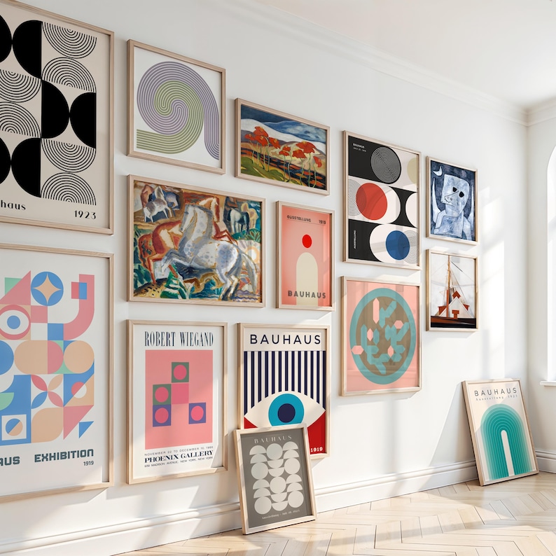 May include: A collection of framed Bauhaus art prints in various sizes and designs, including geometric shapes, abstract compositions, and landscape scenes. The prints feature a range of colors, including black, white, red, blue, and pink. The art is displayed on a white wall.