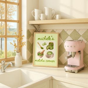 Peut inclure: Une cuisine avec une affiche encadr&eacute;e intitul&eacute;e "michele's matcha corner" pr&eacute;sentant des illustrations de pr&eacute;paration du matcha. Une machine &agrave; expresso rose SMEG est &agrave; c&ocirc;t&eacute; de l'affiche. Des pichets et tasses blancs sont sur une &eacute;tag&egrave;re au-dessus.