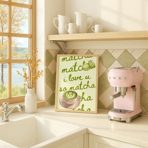 May include: A kitchen interior featuring a framed print with the words "matcha match, I love u so matcha." A pink espresso machine is next to the print. White jugs and cups are on a shelf above. A white vase with yellow flowers is on the counter.