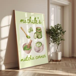 Peut inclure: Une affiche encadr&eacute;e avec des illustrations sur le th&egrave;me du matcha. Elle pr&eacute;sente un bol avec un fouet, une tasse de latte au matcha, des macarons et un pot de poudre de matcha. Le texte "Michele's matcha corner" est en vert.
