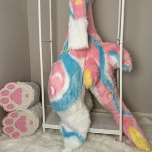 Op de afbeelding: Een volledig lichaamspak van imitatiebont met een levendig patroon van roze, blauwe en witte strepen met gele accenten. Het pak bevat een lange staart en bijpassende witte pootvormige handschoenen met roze kussentjes op de handpalmen.