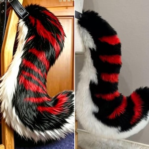 May include: A long, curved faux fur tail featuring a striped pattern of black, vibrant red, and white. The tail is constructed with a black leather-like attachment strap at the top, showcasing a thick, plush texture throughout its entire length.