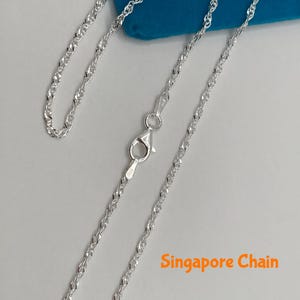May include: A silver Singapore chain necklace with a lobster clasp. The chain is displayed on a light gray surface with a blue background. The text "Singapore Chain" is in orange.