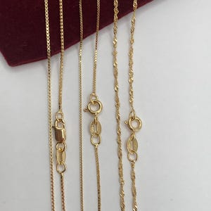 May include: Several gold necklaces with different chain styles are displayed against a white background and a red velvet surface. The necklaces include box chains and twisted rope chains, each with a clasp.