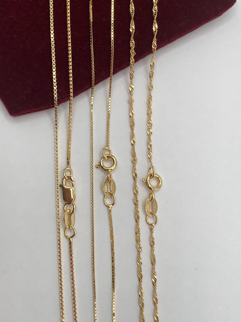 May include: Several gold necklaces with different chain styles are displayed against a white background and a red velvet surface. The necklaces include box chains and twisted rope chains, each with a clasp.