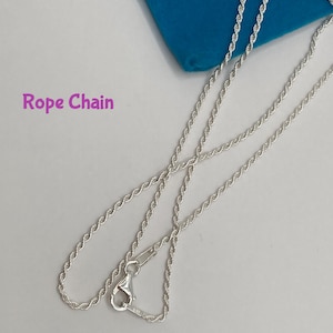 May include: A silver-colored rope chain necklace with a lobster clasp. The chain is displayed on a light gray surface with a blue cloth in the background. The text "Rope Chain" is visible.