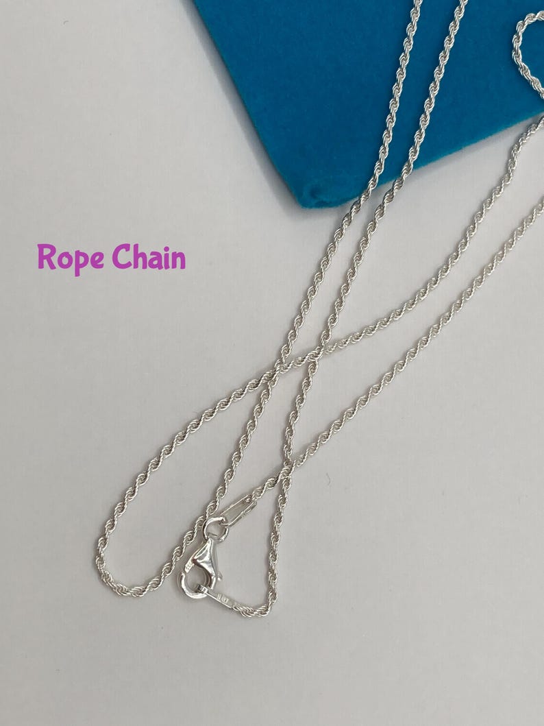 May include: A silver-colored rope chain necklace with a lobster clasp. The chain is displayed on a light gray surface with a blue cloth in the background. The text "Rope Chain" is visible.