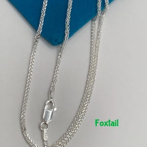 May include: A silver foxtail chain necklace with a lobster clasp. The necklace is displayed on a blue surface with the word "Foxtail" in green text.
