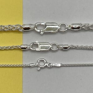 May include: Three silver wheat chains of varying thicknesses arranged horizontally. The top two chains feature lobster claw clasps, while the bottom, thinnest chain is secured with a round spring ring clasp. Each chain has a metallic finish.