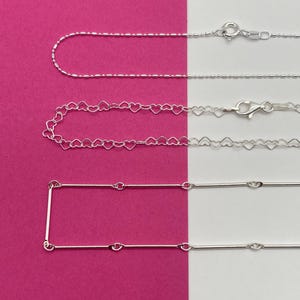 Peut inclure: Trois chaînes en argent présentées horizontalement. La chaîne du haut comporte de petites perles, la chaîne du milieu se compose de maillons en forme de cœur interconnectés, et la chaîne du bas alterne entre fines barres et anneaux.