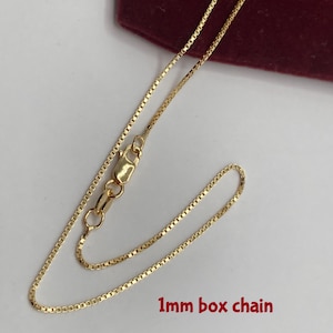 May include: A gold-colored 1mm box chain necklace. The chain is laid out on a white surface with a burgundy background. The chain is a box style with a lobster clasp closure.