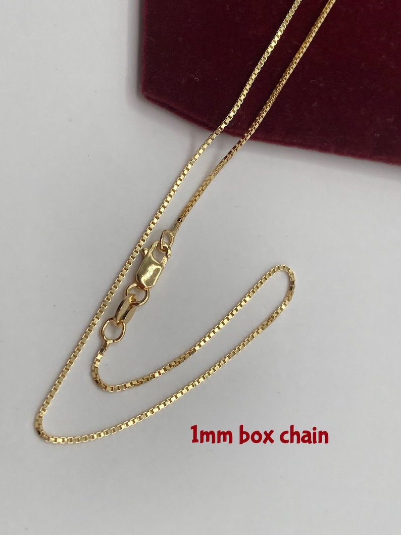 May include: A gold-colored 1mm box chain necklace. The chain is laid out on a white surface with a burgundy background. The chain is a box style with a lobster clasp closure.