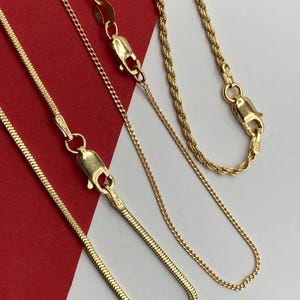 May include: Three gold-toned metal chains of varying styles, including a flat snake chain, a delicate cable chain, and a twisted rope chain. Each chain features a secure lobster claw clasp, with visible metal tags stamped with 925 near the closures.