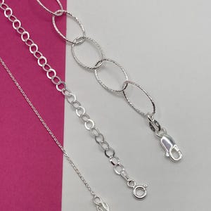 May include: Three silver-toned metal chains of varying styles. One is a fine cable chain, one is a medium-sized round link chain, and one is a large oval link chain with a textured finish. Each chain features a metal clasp at the end.