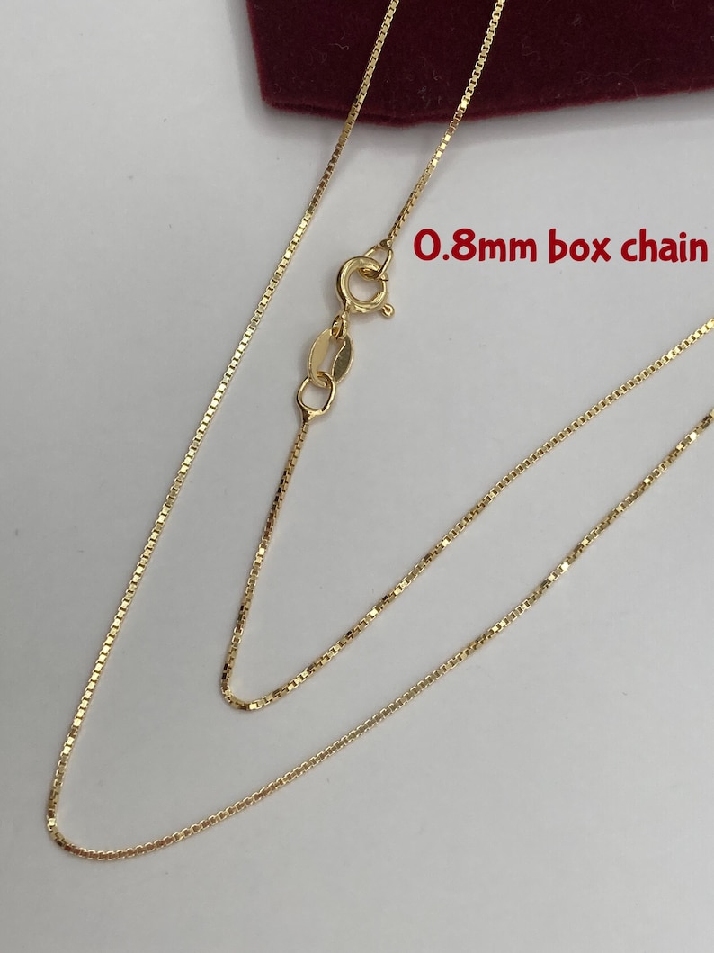 May include: A delicate gold-colored necklace with a box chain design. The chain is 0.8mm thick and features a lobster clasp closure. The necklace is displayed against a neutral background, highlighting its fine details.