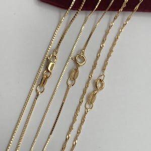 May include: Close-up of several gold necklaces with different chain styles. The chains include box, curb, and twisted rope designs, each with a clasp. The necklaces are displayed on a white surface with a burgundy background.