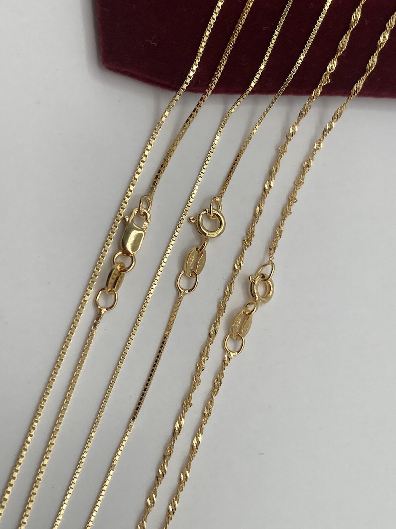 May include: Close-up of several gold necklaces with different chain styles. The chains include box, curb, and twisted rope designs, each with a clasp. The necklaces are displayed on a white surface with a burgundy background.