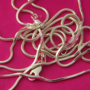 May include: A silver-toned snake chain necklace featuring a smooth, flexible, and rounded link design. The chain is coiled in a loose, overlapping pattern and includes a secure lobster claw clasp mechanism at the end for fastening the jewellery piece.