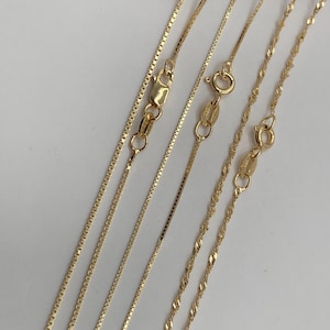 May include: Close-up of several delicate gold necklaces with different chain styles. The chains include box, rope, and twisted designs, each with a secure clasp. The necklaces are displayed against a white background, highlighting their golden color and intricate details.