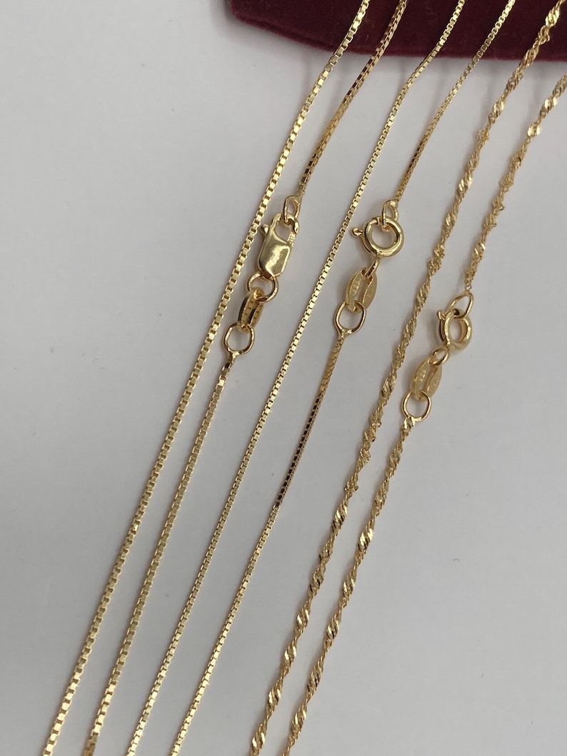 May include: Close-up of several delicate gold necklaces with different chain styles. The chains include box, rope, and twisted designs, each with a secure clasp. The necklaces are displayed against a white background, highlighting their golden color and intricate details.