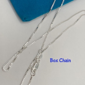 May include: A silver box chain necklace with a lobster clasp, displayed on a white surface with a blue cloth in the background. The text "Box Chain" is visible in the image.