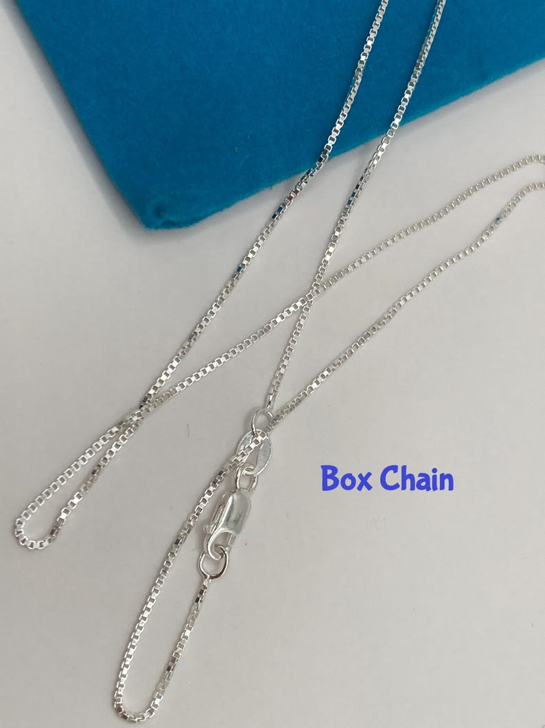 May include: A silver box chain necklace with a lobster clasp, displayed on a white surface with a blue cloth in the background. The text "Box Chain" is visible in the image.
