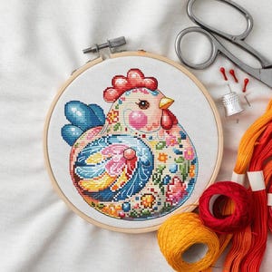 May include: A colorful cross-stitch of a chicken in a wooden embroidery hoop. The chicken features a red comb, pink cheeks, and a patterned design with blue, pink, yellow, and floral elements. Embroidery supplies, including scissors and thread, are visible.