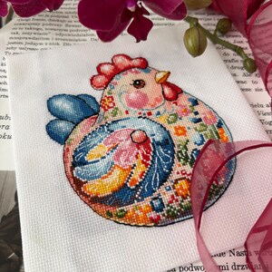 May include: A colorful cross-stitch of a chicken with a red comb, blue wings, and a patterned body. The embroidery is on white fabric, with a background of a book and purple orchids.