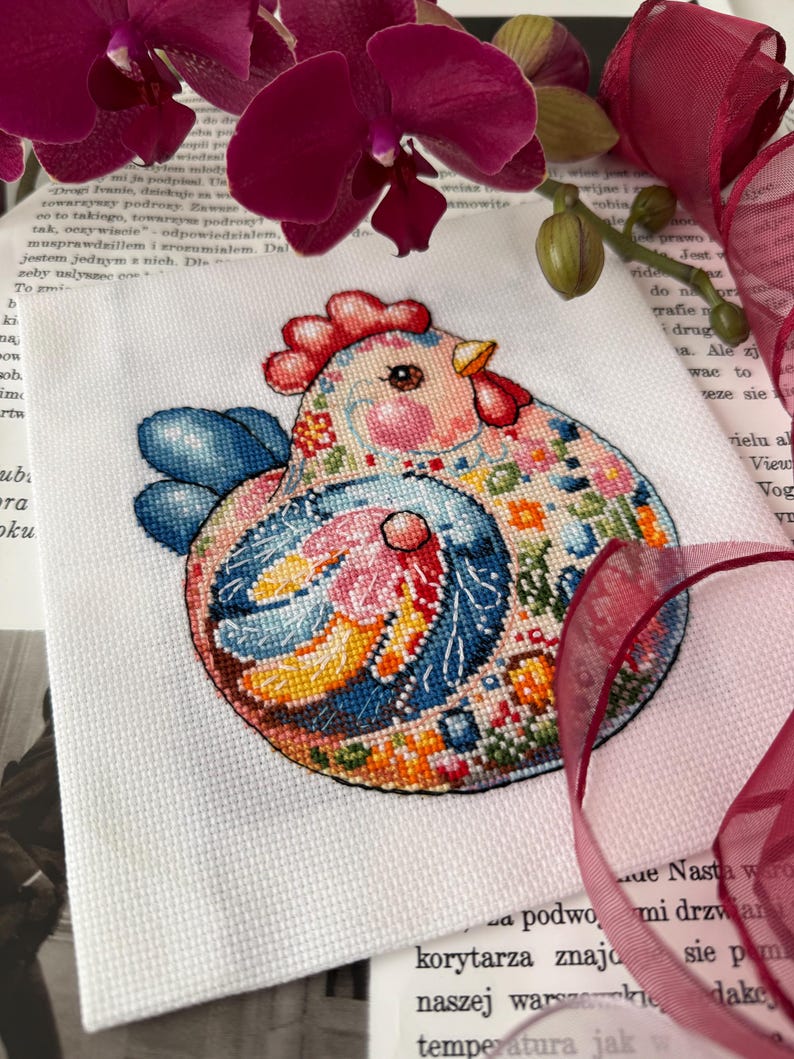 May include: A colorful cross-stitch of a chicken with a red comb, blue wings, and a patterned body. The embroidery is on white fabric, with a background of a book and purple orchids.