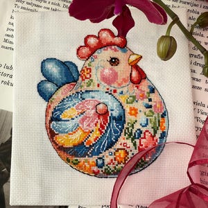 Op de afbeelding: Een kleurrijk kruissteekpatroon van een kip met bloemmotieven. De kip heeft een rode kam en lellen, blauwe vleugels en staat tegen een witte achtergrond. Het ontwerp bevat roze, oranje en blauw.