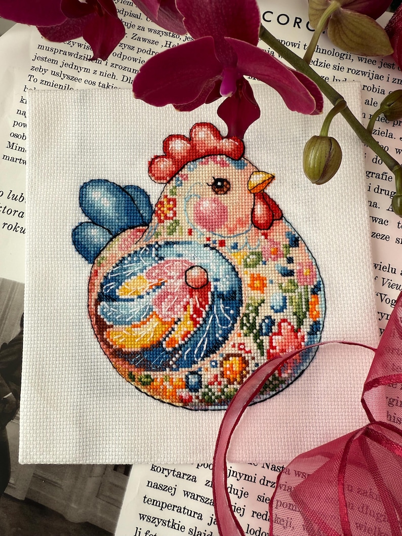 May include: A colorful cross-stitch of a chicken with floral patterns. The chicken has a red comb and wattle, blue wings, and is set against a white background. The design incorporates various colors, including pink, orange, and blue.