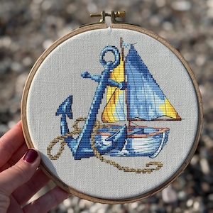 Peut inclure: Broderie au point de croix d'un voilier et d'une ancre dans un cercle en bois. Le voilier a une voile bleue et une voile jaune. L'ancre est bleue et est reliée au bateau par une corde marron. Le fond est un tissu gris clair.